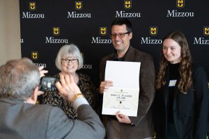Three individuals smiling while posing for a photo. One of the individuals is holding an award certificate. There is a person on the left taking a photo of the three individuals with a cell phone.