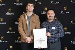 Two individuals smiling while posing for a photo. One of the individuals is holding an award certificate.