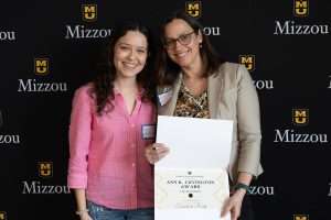 Two individuals smiling while posing for a photo. One of the individuals is holding an award certificate.